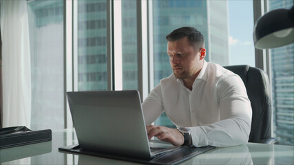 Businessman behind a laptop in the office. A businessman in a white shirt sits at a table in a skyscraper.