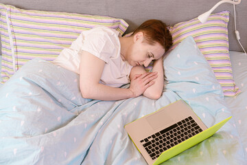 Young red-haired woman fell asleep in bed while watching a series. The woman fell asleep in front of an open laptop. Workaholism, a lot of work, addiction to gadgets. The woman is sleeping in bed.