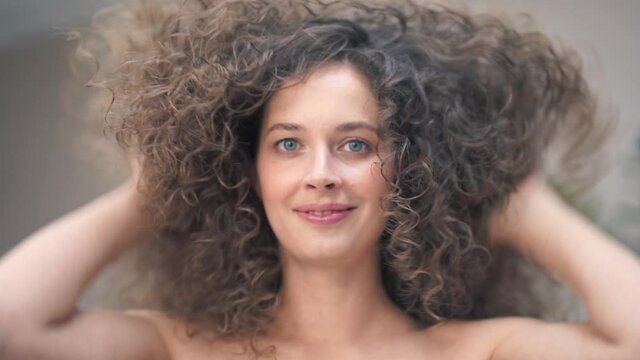 Close-up of a beautiful young woman with curly hair, she tilts her head in different directions, smiling at the camera, touching her hair with her hands. High quality 4k footage