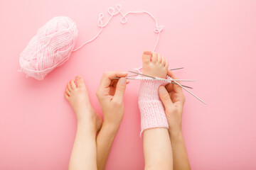 Young adult mother try on unfinished wool knitted sock on her daughter little foot. Pink table...