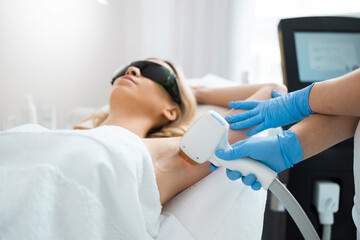 Woman on underarm laser epilation procedure in beauty clinic. Beautician removing hair of pretty...