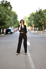 A beautiful young girl in a black suit and sunglasses walks around the city.