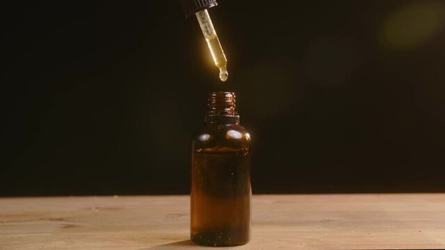 Handheld shot of a hand, filling a small glass dropper bottle with CBD hemp oil, on the black background.