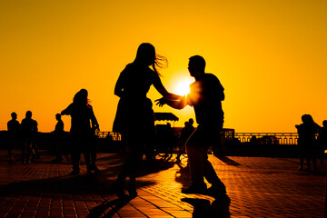 Fototapeta na wymiar Woman and man couple silhouettes dancing against warm sunset orange sky on quay at evening - sun lens flare. Group dance, romantic, love, summer outdoor activity and vacation concept