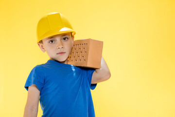 Boy in a construction helmet holds a brick in his hands on yellow background