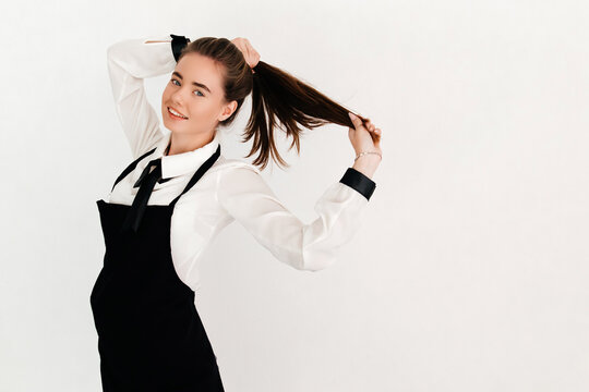 Portrait of a confident waitress with a ponytail, wearing white short, black apron and tie, fixing her hair before welcoming guests, isolated on white background