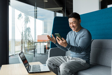 Successful Asian businessman celebrates victory, looks at phone, happy and rejoices sitting in...