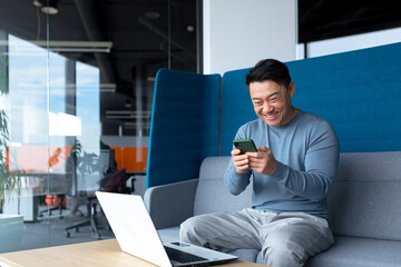 Successful Asian businessman celebrates victory, looks at phone, happy and rejoices sitting in modern office