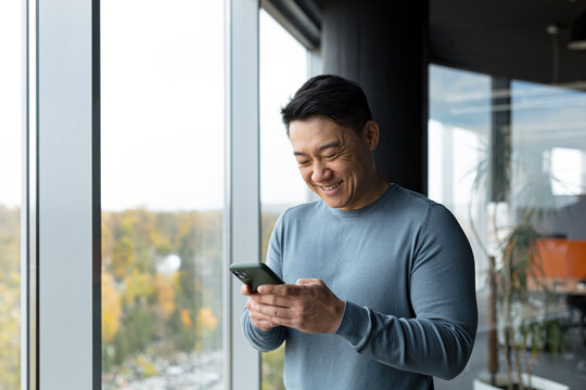 Successful and happy Asian man uses the phone online in the office by the window