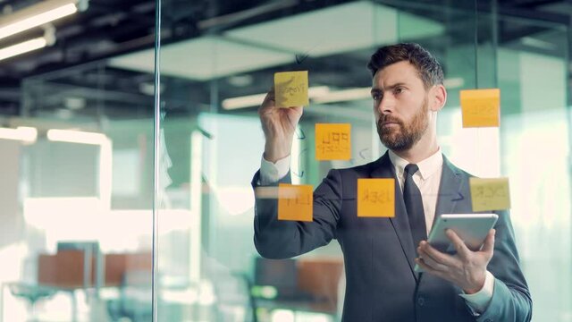 serious businessman analyst develops a strategy for enterprise, financial growth thinking make startup plan in office Business male employee in suit write on paper sticker sticky notes on glass wall