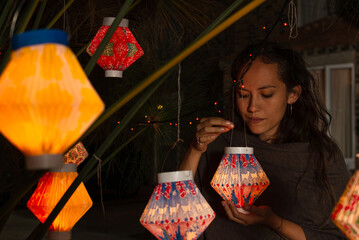 Hispanic woman holding a paper lantern while looking at it, under a plant decorated with multiple lanterns - obrazy, fototapety, plakaty