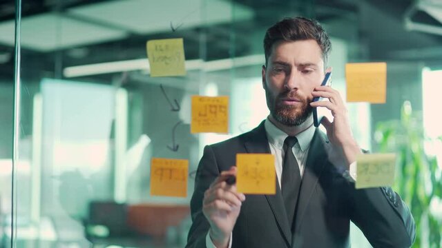 business man talking on the phone in a modern office behind a glass task partition wall with paper stacks checking information on ideas board hanging plans on colored paper stickers, plans for future