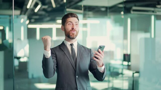 Happy euphoric bearded business man wear suit celebrate mobile win, excited overjoyed lucky executive winner receive good news in message use smartphone rejoice bet financial success victory in office