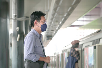 Asian man commuter with face mask on standing on the subway platform waiting to go inside train. Masked transit concept.

