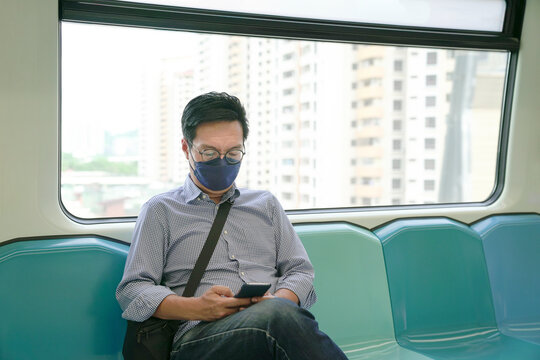 Asian man commuter wearing face mask looking at his smartphone, sitting inside a subway moving train. Masked transit concept.
