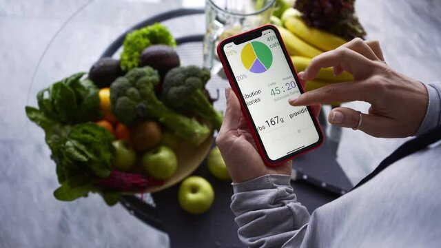 Unrecognizable lady uses application on smartphone to check calories of recipe on table with fresh fruits and vegetables in contemporary kitchen close view