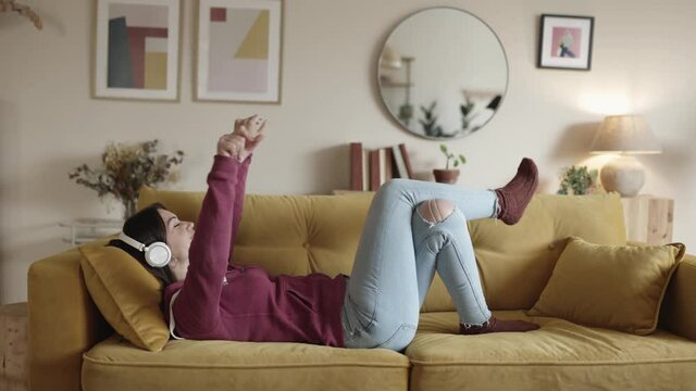 Side view of young happy woman with closed eyes lying on sofa and listening to music in wireless headphones and dancing. High quality 4k footage