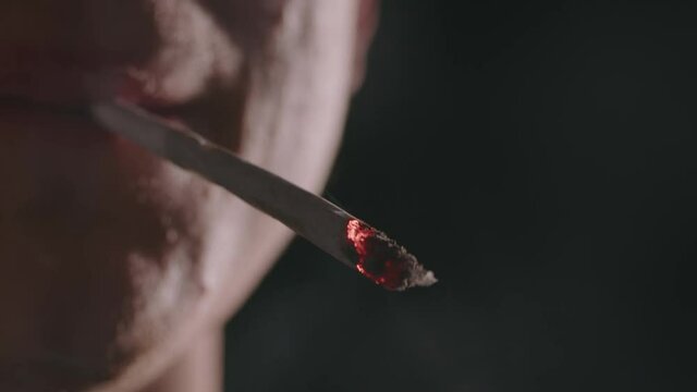 Close-up shot of a lit marijuana joint. Man smoking weed cigarette facing camera.
