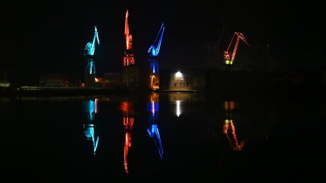 Cranes illuminated with colorful lights in shipyard. Harbour in Adriatic sea at night. Jackup rig, Oil platform and Container crane in Dockyard. Ship port. Industry and transport. Shipbuilding.  