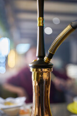 Hookah on table in lounge bar. Close-up of black pipe and golden shisha flask. Smoking person in blurry background. Leisure in oriental cafe.