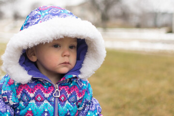 18 month old baby girl in a colorful patterned snow suit and hood looks away from the camera with a serious expression; waiting for the snow to fall