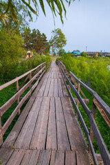 Fototapeta premium Wooden bridge in the village across the river, near grass, bushes and trees.