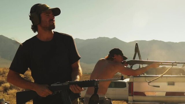 Two young men shooting guns for fun in the desert. Static medium shot.