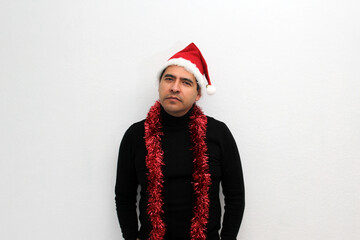 Dark-haired Latin adult man with hat and Christmas garland shows his anger, disgust and sadness for the arrival of December, he does not like Christmas
