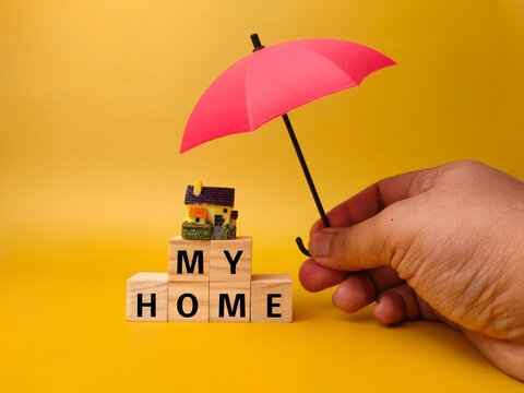 Hand holding red umbrella,miniature house and wooden cube with word MY HOME on a yellow background. Business concept.