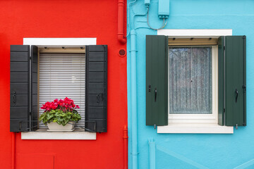 Close up of a brightly colored plaster facade with two windows and a flower pot, the leftmost half is red and the rightmost is light-blue