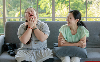 Asian young chubby down syndrome autistic autism little cute daughter sitting on cozy sofa smiling laughing having good time and fun with lovely mother after playing video console game hobby together