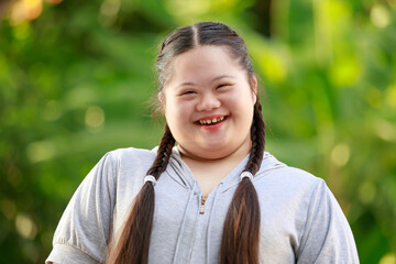 Portrait shot of Asian young chubby down syndrome autistic autism little cute schoolgirl with braid pigtail hairstyle model stand posing smiling look at camera in front blurred garden view background - obrazy, fototapety, plakaty