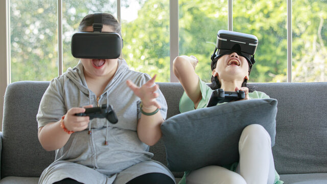 Asian happy lovely mother and young chubby down syndrome autistic autism little daughter sitting on cozy sofa wearing VR virtual reality goggles smiling holding joysticks playing console video game