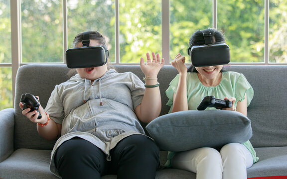Asian happy lovely mother and young chubby down syndrome autistic autism little daughter sitting on cozy sofa wearing VR virtual reality goggles smiling holding joysticks playing console video game