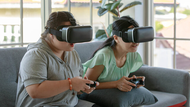 Asian happy lovely mother and young chubby down syndrome autistic autism little daughter sitting on cozy sofa wearing VR virtual reality goggles smiling holding joysticks playing console video game