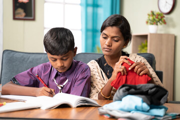 Focus on son, Mother helping her child to do homework while busy folding cloths at home - concept...