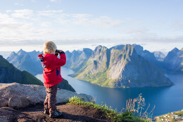 Cute child, standing on top of the mountains and looking down on Reine after climbing Reinebringen treeking path with lots of stairs, using binoculars, Lofoten