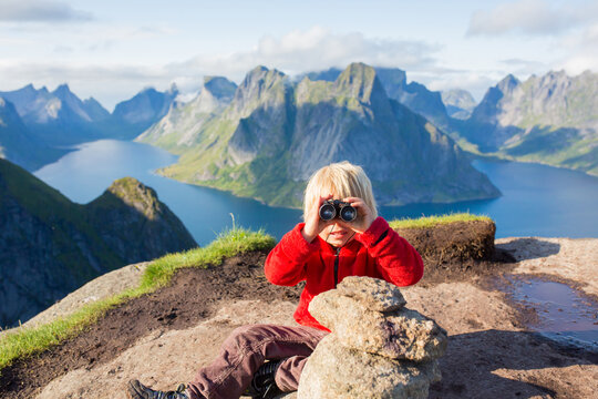 Cute child, standing on top of the mountains and looking down on Reine after climbing Reinebringen treeking path with lots of stairs, using binoculars