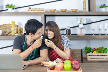 Happy young Asian healthy couple with casual clothes is smiling and feeding fresh fruit each other in kitchen at home in holiday