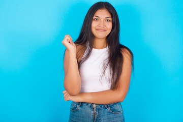 Fototapeta na wymiar Cheerful Young hispanic girl wearing tank top over blue background with hand near face. Looking with glad expression at the camera after listening to good news. Confidence.