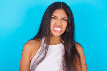 Mad crazy Young hispanic girl wearing tank top over blue background clenches teeth angrily, being...