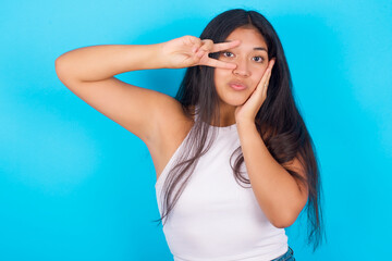 Young hispanic girl wearing tank top over blue background making v-sign near eyes. Leisure lifestyle people person celebrate flirt coquettish concept.