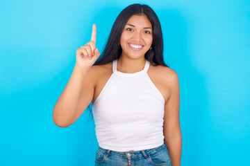 Fototapeta premium Young hispanic girl wearing tank top over blue background showing and pointing up with fingers number one while smiling confident and happy.