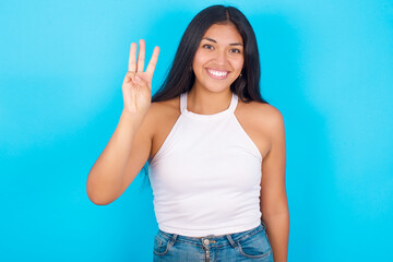 Young hispanic girl wearing tank top over blue background showing and pointing up with fingers number three while smiling confident and happy.