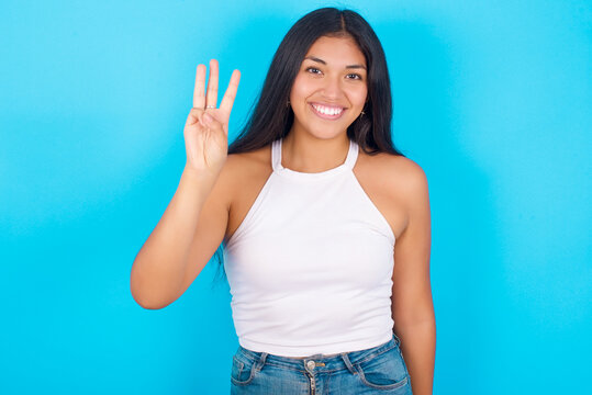 Young Hispanic Girl Wearing Tank Top Over Blue Background Showing And Pointing Up With Fingers Number Three While Smiling Confident And Happy.