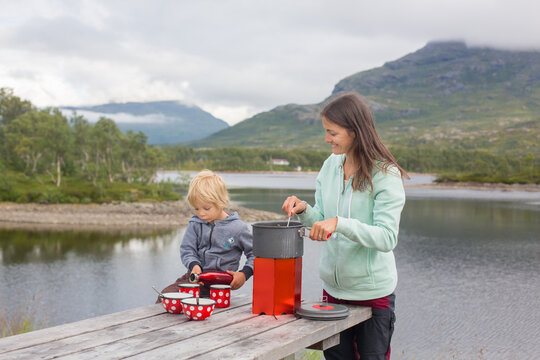 Child, Eating On A Bench On A Rest Stop Along The Road, Mom Cooking On Small Camping Cooker And Having Family Lunch On The Road