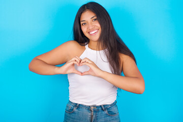 Young hispanic girl wearing tank top over blue background smiling in love showing heart symbol and shape with hands. Romantic concept.