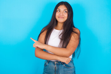Fototapeta na wymiar Portrait of Young hispanic girl wearing tank top over blue background posing on camera with tricky look, presenting product with index finger. Advertisement concept.