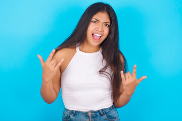 Fototapeta premium Born to rock this world. Joyful Young hispanic girl wearing tank top over blue background screaming out loud and showing with raised arms horns or rock gesture.