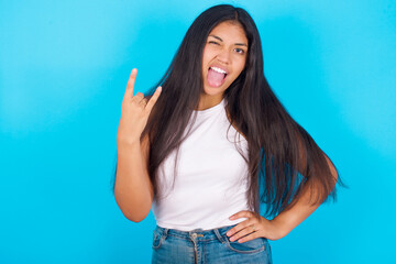 Fototapeta na wymiar Portrait of a crazy Young hispanic girl wearing tank top over blue background showing tongue horns up gesture, expressing excitement of being on concert of band.
